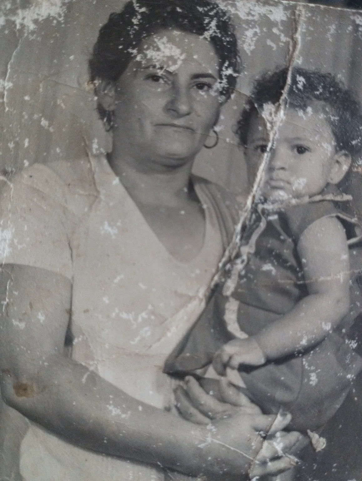 Fotografia antiga de uma mãe segurando um bebê, coberta de manchas brancas, rasgos e marcas do tempo.